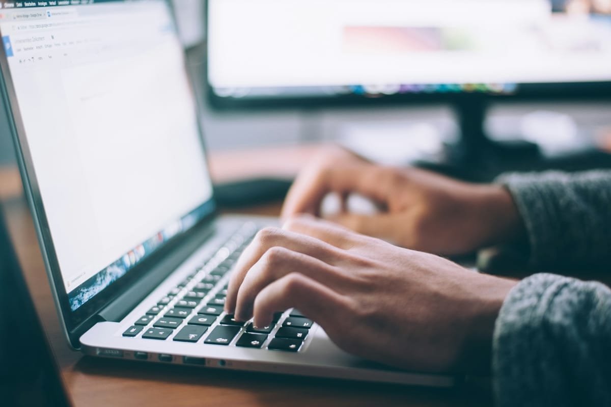 Manos escribiendo en una laptop sobre un escritorio de madera, con un monitor al fondo.